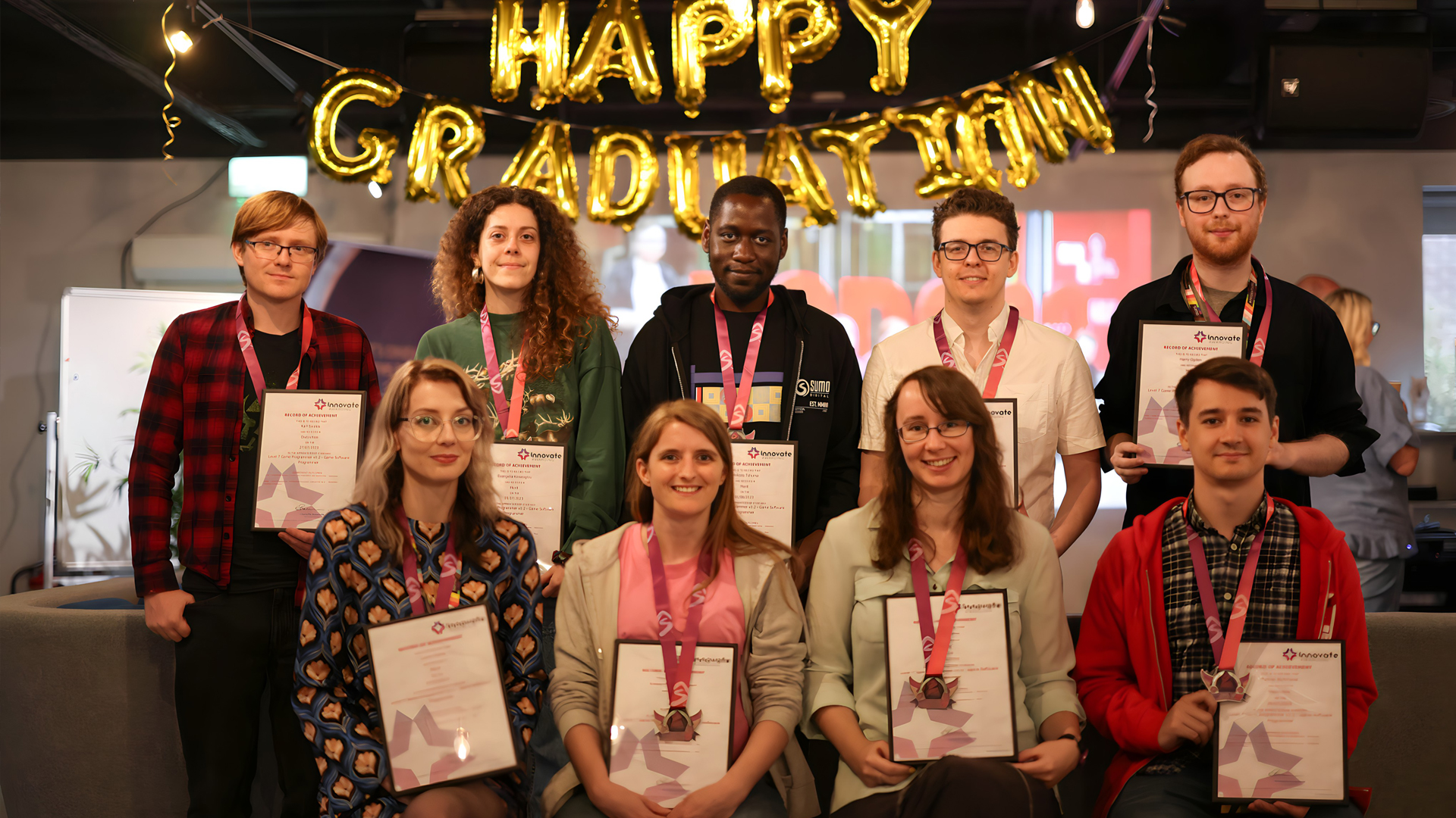 A group of nine people pose indoors, holding certificates and wearing medals. Behind them, gold balloon letters spell HAPPY GRADUATION. The mood is celebratory and cheerful.