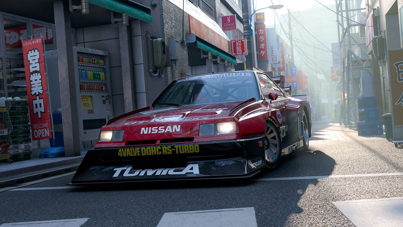 A red Nissan Skyline racing car with black detailing and a large front splitter is parked on a sunlit city street lined with shops and Japanese signs.