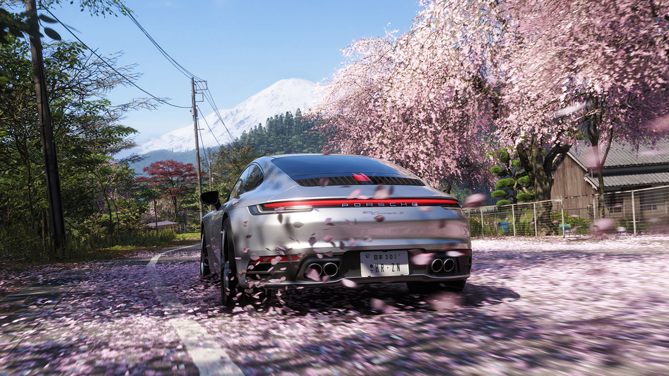 A silver Porsche drives down a road lined with cherry blossom trees in full bloom. Pink petals fill the air and cover the ground, with a snow-capped mountain visible in the background.