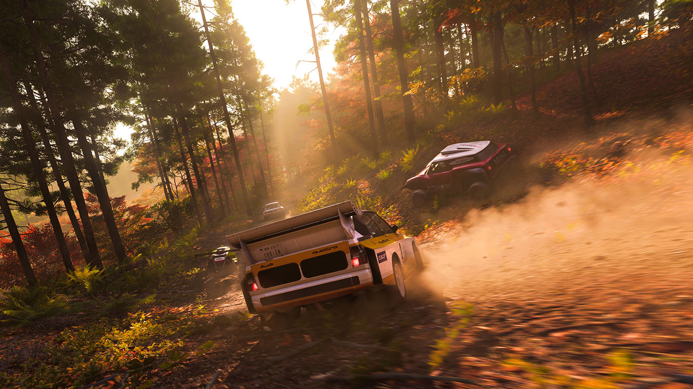 A group of rally cars race through a sunlit, forested dirt road, kicking up dust as they speed round a curve surrounded by tall trees and autumn foliage.