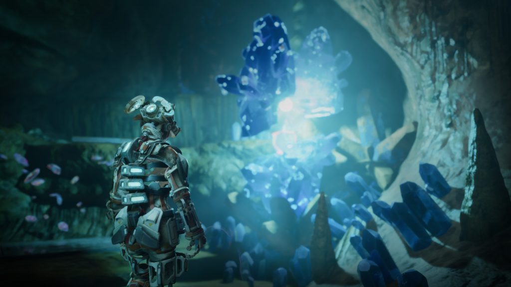 A person in a futuristic, armoured suit explores a cave filled with glowing blue crystals, concepting the scene as one large, radiant crystal illumines the underground environment.