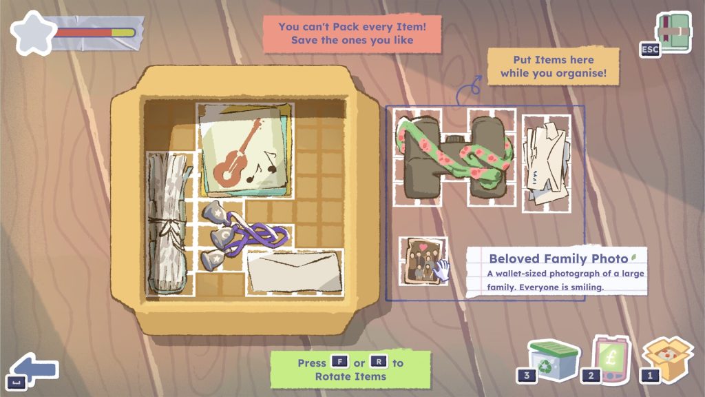 A screenshot from A Storied Life: Tabitha showcasing the packing screen, featuring a cardboard box atop a wooden table. There is a grid inside the box, presenting a puzzle to the player about fitting in different sized items into the limited space available. There are items arranged inside the box and some still outside. A hand icon hovers over one of these items outside the box, bringing up text on a lined piece of paper that reads 'Beloved Family Photo. A wallet-sized photograph of a large family. Everyone is smiling.' Other text on this screen reads 'You can't pack every item. Save the ones you like. Put items here while you organise. Press F or R to rotate items.'