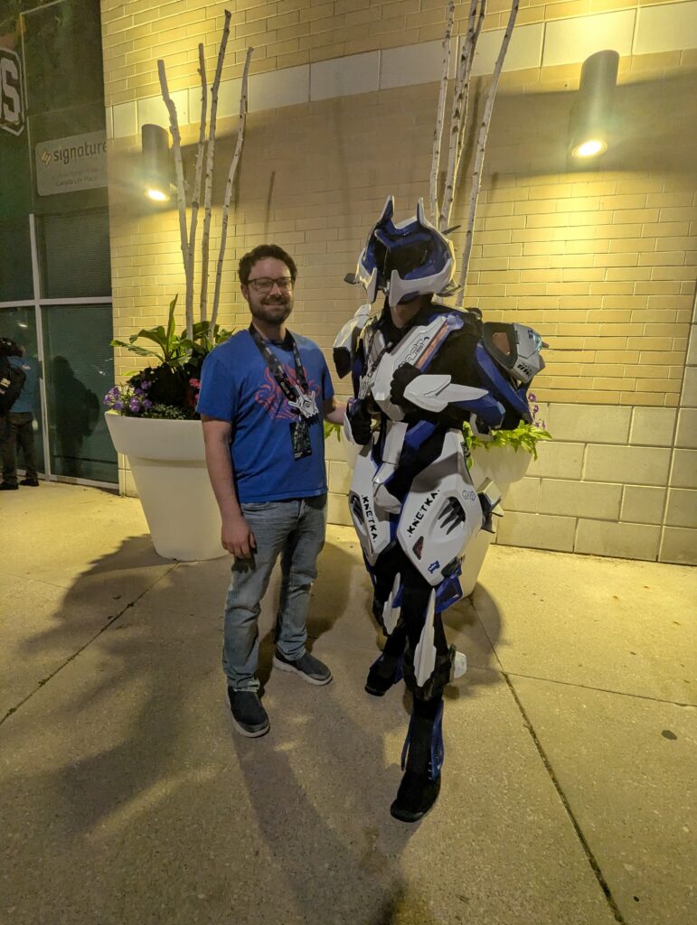 Stephen Gibson and Gauss Moto Cosplayer BagaProps at TennoCon 2026. The photo shows two people stood side by side in a warmly lit outside nighttime environment by the side of a building. The person on the left wears and blue shirt and some jeans, whereas the person to the right is cosplaying Gauss Moto and is in a full robotic suit.